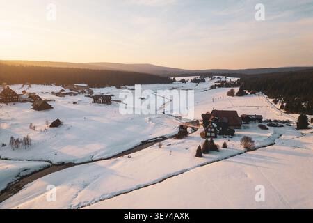 Das Bergdorf Jizerka zeigt im Winter Häuser und schneebedeckte Felder. Die Luftaufnahme fängt die Landschaft mit Bäumen und einem Fluss bei Sonnenuntergang ein. Stockfoto