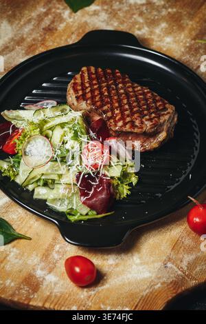 Gegrilltes Gourmet-Rindersteak mit perfekten Saibling-Markierungen, serviert mit einem frischen Gartensalat aus Gurken-Kirschtomaten und Parmesan auf einer schwarzen Grillpfanne. Stockfoto