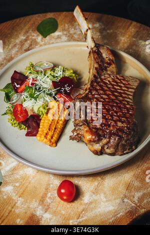Gegrilltes Tomahawk-Ribeye-Steak mit Saibensaibspuren, serviert mit gegrilltem Mais, frischem Gartensalat und Kirschtomaten auf einer Keramikplatte. Stockfoto