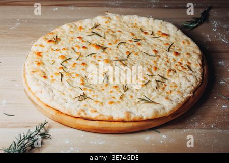 Traditionelles italienisches Focaccia-Brot mit geschmolzenem Käse und frischen Rosmarinnadeln, serviert auf einem rustikalen Holzbrett mit Mehl. Stockfoto