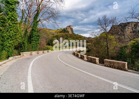 Eine gekrümmte Bergstraße führt in Richtung eines Meteora-Klosters, das auf einer Felssäule in Griechenland thront. Dramatische Wolken und natürliches Grün schaffen eine malerische trave Stockfoto