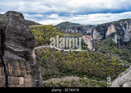 Panoramablick auf Meteora Felsformationen mit dem Kloster Roussanou auf einer schmalen Klippe. Eine kurvige Straße und ein bewaldetes Tal unterhalb der ddr verstärken die ddr Stockfoto