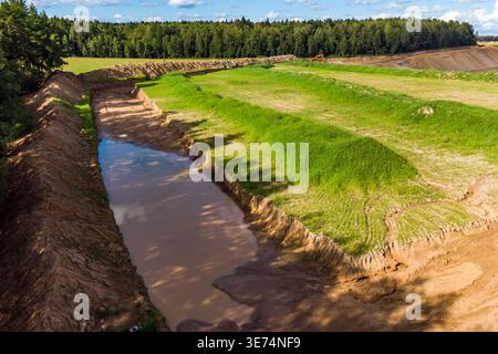 Ausgegrabener Erdkanal mit schlammigem Wasser und üppig grünen Grasbänken, von oben betrachtet. Weit entfernter Wald und ein einzelner Bagger deuten auf laufende Arbeiten hin. Stockfoto