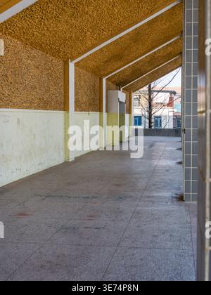 Leerer überdachter Außenkorridor mit einer strukturierten gelben Decke, gefliestem Boden und Stützsäulen, die von natürlichem Sonnenlicht beleuchtet werden. Die Szene wird erneut angezeigt Stockfoto