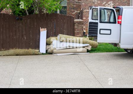 Weißer Van sitzt auf einer Betoneinfahrt. Seine Tür ist offen und offenbart ein dunkles Inneres. In der Nähe des Zauns stapeln sich grünes Gras und Schutzmaterialien. Stockfoto