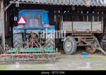 Ein verwitterter blauer Traktor und ein hölzerner Anhänger sitzen in einer rustikalen Scheune zwischen landwirtschaftlichen Arbeitsgeräten und Werkzeugen. Ein braunes Huhn peckt neben einer Wasserschale, was an ländliche L Stockfoto