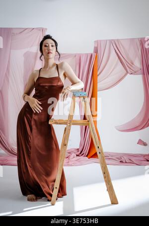 Erwachsene asiatische Chinesin posiert im Studio, lehnt sich auf Holzleiter mit Satinkleid und Vorhängen. Mode, Eleganz, minimalistisch, Lookbook, Hintergrund, Stockfoto