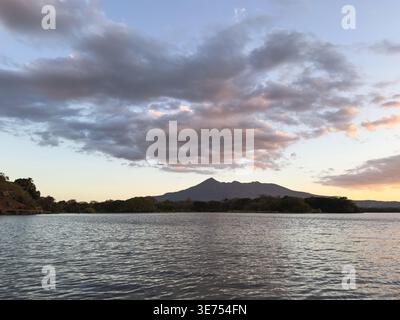 Das Wasser reflektiert den farbenfrohen Himmel, während die Sonne hinter dem Berg untergeht und eine malerische Aussicht bietet. Bäume säumen das Ufer mit ruhigem Wasser. Stockfoto