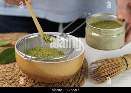 Frau, die Matcha-Pulver in eine Schüssel am weißen Holztisch siebt, Nahaufnahme Stockfoto