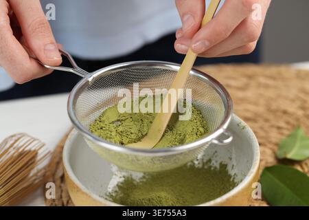 Frau, die Matcha-Pulver in eine Schüssel am weißen Tisch siebt, Nahaufnahme Stockfoto