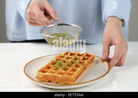 Frau siebt Matcha-Pulver auf Waffeln an weißem Holztisch, Nahaufnahme Stockfoto