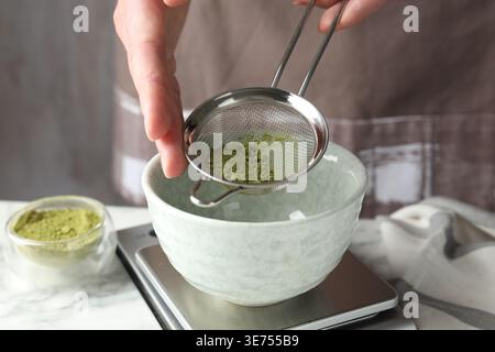 Frau, die Matcha-Pulver in eine Schüssel am weißen Tisch siebt, Nahaufnahme Stockfoto