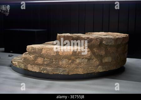 Eine Nahaufnahme einer alten runden Backstein- und Steinstruktur, die im Inneren auf einer schwarzen runden Plattform ausgestellt ist. Das verwitterte Mauerwerk zeigt mehrere Stockfoto