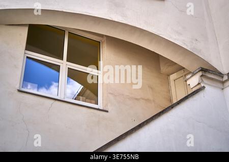 Eine nach oben gerichtete Ansicht eines architektonischen Details mit einem geschwungenen Torbogen an einer verwitterten beigen Stuckfassade. Ein weiß gerahmtes Fenster mit Stockfoto