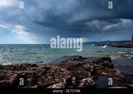 Dramatische Sturmwolken über Rocky Shoreline mit entferntem Leuchtturm und abgehacktem Wasser Stockfoto