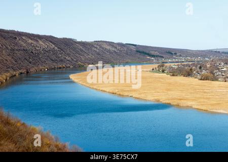 Ein ruhiger Fluss fließt durch ein Tal, das von Hügeln umgeben ist. Das Wasser ist klar und blau, mit grasbewachsenen Ufern und einer entfernten Siedlung Stockfoto