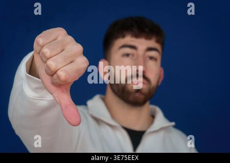 Fokussierter junger Mann, der Abneigung und Unzufriedenheit mit einer negativen Geste mit Daumen nach unten ausdrückt, was auf Ablehnung oder Misserfolg hinweist Stockfoto