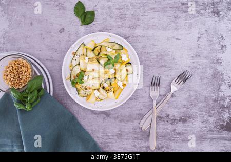 Blick von oben auf eingelegten Zucchini-Salat mit Fetakäse, Pinienkernen und Basilikum auf grauer Oberfläche mit Besteck Stockfoto