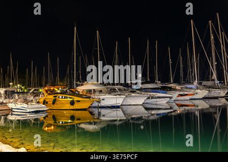 Nachtjachthafen Pier zwischen verankerten Yachten unter dunklem Sternenhimmel, die ruhiges Wasser reflektieren. Hafendock Perspektive Boote Masten Seile Stockfoto