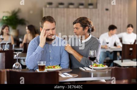 Zwei Freunde treffen sich in einem modernen Restaurant - der Mann tröstet seinen verärgerten, traurigen Freund Stockfoto