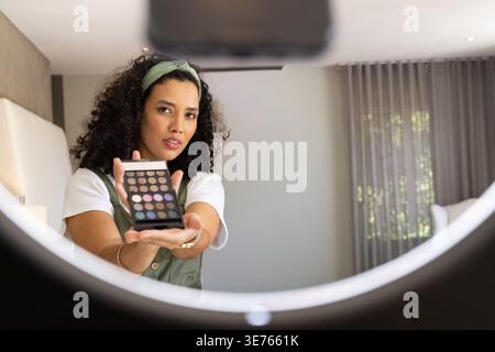 Frau mit grünem Stirnband, das Lidschattenpalette hält und Schatten im Schlafzimmer mit Ringlicht zeigt. Make-up, Kosmetik, Schönheit, Tutorial, Influencer, m Stockfoto