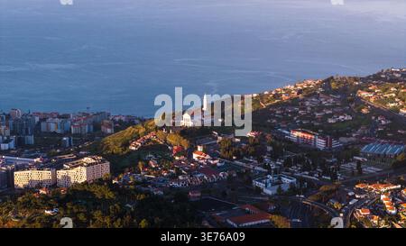 Lufthügelviertel in der Nähe von Funchal, Madeira Island, mit einem weißen Kirchturm, Terrassenhäusern, gewundenen Straßen und ruhigem Meer zur goldenen Stunde Stockfoto