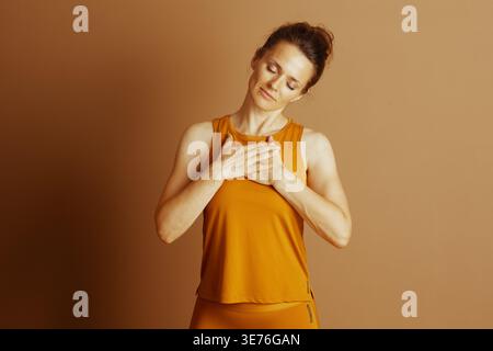 Eine Frau in orangefarbener Activewear mit geschlossenen Augen und gefalteten Händen steht in einer ruhigen, achtsamen Pose. Die sanfte Beleuchtung verstärkt die ruhige, friedliche Stimmung, die an Sie erinnert Stockfoto