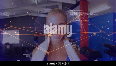 Lächelnde blonde Frau, die im Fitnessstudio steht, Tanktop und weißes Handtuch trägt und digitale Überzüge zeigt Stockfoto