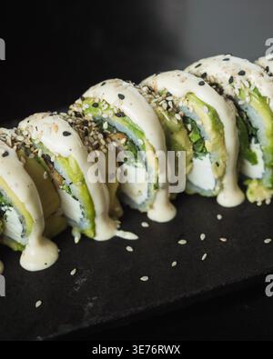 Frische Sushi-Brötchen mit Avocado, Sesam und cremiger Sauce. Die schwarze Schieferplatte sorgt für eine elegante Präsentation. Sesamsamen sorgen für Textur und visuellen Kontrast. Stockfoto