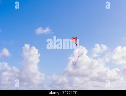 Buntes, an einem dünnen Faden befestigtes Luftspielzeug schwingt über eine Wolkenlandschaft. Die Weitwinkelaufnahme bildet den fliegenden Drachen vor einem tiefblauen Himmel. Stockfoto