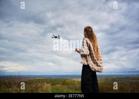 Rückansicht einer Frau, die Drohne kontrolliert, die bei bewölktem Himmel über weite Landschaft fliegt. Pilotin mit Fernbedienung auf dem Hügel, Luftaufnahmen und Explorationskonzept der Technologie Stockfoto