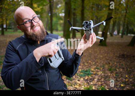 Der Mann zeigt auf die Drohne und zeigt sie der Kamera. Ein lächelnder bärtiger Mann, der Quadcopter und Controller in einem Park hält und Ausrüstung für Luftfotografie, Konzept moderner Technologie und Hobby präsentiert Stockfoto