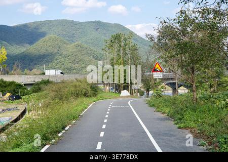 Ein schmaler Radweg führt durch die Vegetation am Fluss in Ulju-Gun, mit Berghängen, einem Brückenanstieg und einem Warnschild am Straßenrand. Stockfoto