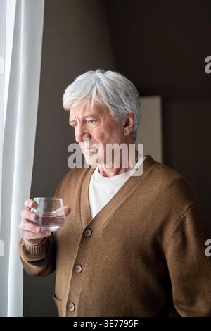 Trauriger, trauriger alter Mann, der Wasser aus Glas trinkt und aus dem Fenster blickt. Einsamkeit und Alterskrankheit Stockfoto