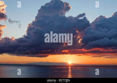 Atemberaubender Sonnenuntergang über dem Meer mit großen dunklen Wolken, Bergsilhouetten und goldenen Sonnenspiegeln auf dem Wasser Stockfoto