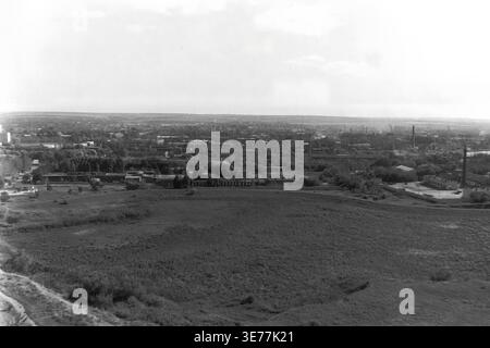 Sloviansk, 1997, aus dem SKTB-Gebäude. Ein breiter, erhöhter Panoramablick auf das Schwarzweiß-Archiv mit Blick auf ein ausgedehntes, offenes Feld in Richtung eines entfernten Industriegebiets und Wohnviertels. Das weitläufige postsowjetische Stadtbild zeigt hohe Fabrikschornsteine, die über den dichten Baumdächern ragen und die typische Integration von Industrie und Wohnraum veranschaulichen. Dieses authentische Vintage-Foto zeigt das weitläufige Stadtbild und die alltägliche städtische Umgebung in einem blühenden, friedlichen Donbass. Stockfoto