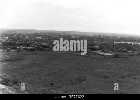 Sloviansk, 1997, aus dem SKTB-Gebäude. Ein breiter, erhöhter Panoramablick auf das Schwarzweiß-Archiv mit Blick auf ein ausgedehntes, offenes Feld in Richtung eines entfernten Industriegebiets und Wohnviertels. Das weitläufige postsowjetische Stadtbild zeigt hohe Fabrikschornsteine, die über den dichten Baumdächern ragen und die typische Integration von Industrie und Wohnraum veranschaulichen. Dieses authentische Vintage-Foto zeigt das weitläufige Stadtbild und die alltägliche städtische Umgebung in einem blühenden, friedlichen Donbass. Stockfoto