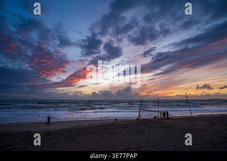 Ein spektakulärer feuriger Sonnenuntergang beleuchtet die Wolken in Rot und Orange über einem einsamen Strand. Im Vordergrund ein provisorisches Fußballtor und Silhouetten von Stockfoto