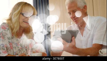 Lächelndes Seniorenpaar, das sich ein Tablet in einer Essecke teilt, in einer Bluse mit Blumenmuster, weißem Hemd, Gläsern, Tassen Stockfoto