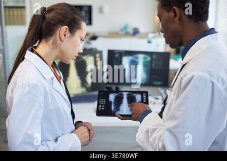 Kaukasische junge Erwachsene Frau und schwarzer junger erwachsener Mann, die in der Klinik Thoraxröntgenaufnahmen auf Tablette überprüfen, Diagnose und Teamarbeit diskutieren. Nützlich für die Zusammenarbeit im Gesundheitswesen und das Radiologiemarketing Stockfoto