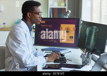 Schwarzer männlicher Arzt, der digitale Patientenakten und Thoraxscans auf Desktop-Monitoren überprüft und so den telemedizinischen Arbeitsablauf unterstützt. Ideal für Gesundheitstechnologie, Diagnostik und medizinische Datenthemen Stockfoto