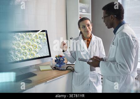 Kaukasische Frau und afroamerikanische männliche Wissenschaftler diskutieren im Labor über Zellanalyse neben Desktop-Monitor und Tablet. Ideal für Biotech-Forschung und Innovationsmarketing im Gesundheitswesen Stockfoto