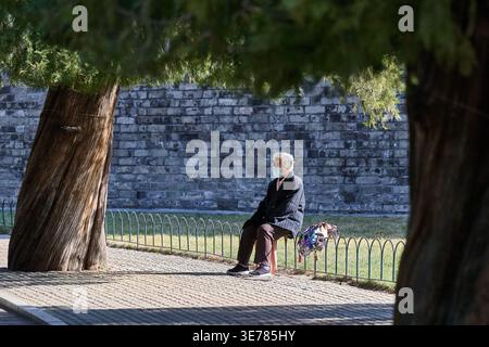 Senior Mann mit medizinischer Gesichtsmaske, der auf einem Hocker vor einer Steinmauer in einem öffentlichen Park sitzt, Peking, China, Asien. Stockfoto