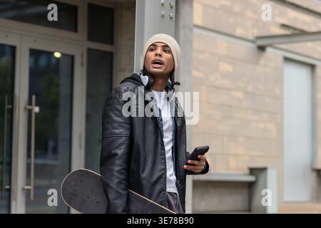 Mann, der sich am Eingang gegen die Säule lehnt, trägt eine Mütze, die Skateboard hält und das Smartphone überprüft. Urban, Outdoor, Lifestyle, Kopfhörer, Telefon, Skate, jac Stockfoto