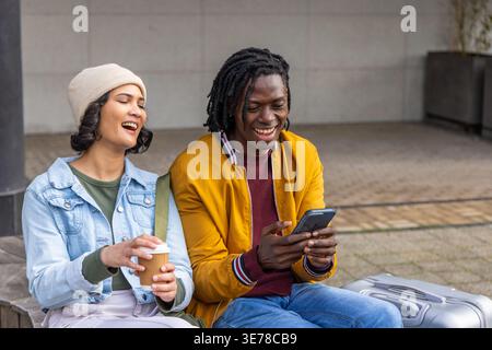 Diverse Paare sitzen auf einer niedrigen Bank am plaza, eine Frau hält Kaffee, ein Mann mit Smartphone lächelnd Stockfoto