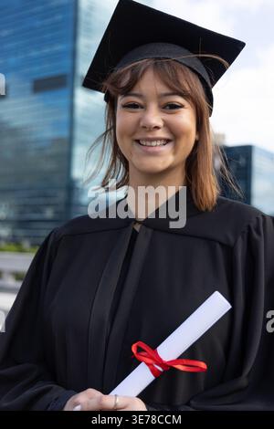 Asiatische Frau lächelt mit gerolltem Diplom mit rotem Band und schwarzer Mütze in der Nähe von Glastürmen Stockfoto
