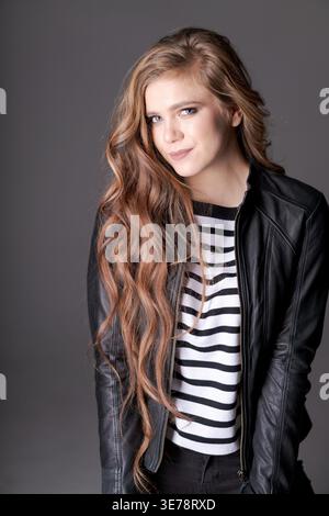 Eine Frau mit langen braunen Haaren trägt eine schwarze Lederjacke und ein weiß gestreiftes Hemd. Sie lächelt und posiert für die Kamera Stockfoto