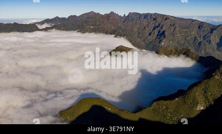 Luftbild zeigt Pico Ruivo und Basaltkämme über niedrigen Wolken auf Madeira, Morgenlicht wirft lange Schatten auf Terrassenhänge, weite Panoramasicht, Stockfoto