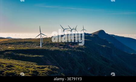 Aus der Vogelperspektive eines Windparks auf Madeira Island, Portugal, mit weißen Turbinen auf mit Sträuchern bedeckten Bergen, einem markanten Gipfel und einem Wolkenmeer Stockfoto