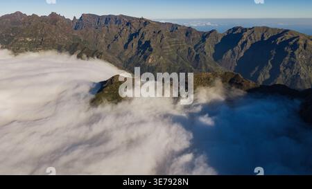 Drohnenblick auf die zentralen Berge Madeiras, Pico do Areeiro nach Pico Ruivo, schiere Basaltklippen über sanften Wolken, Morgenlicht, tiefe Schatten, Stockfoto